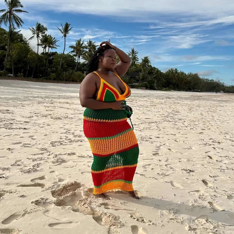 Mulher com saída de praia de crochê nas cores verde, amarelo e vermelho posa em uma praia de areia clara. Palmeiras ao fundo. Ela usa o cabelo preso, com uma das mãos na cabeça.