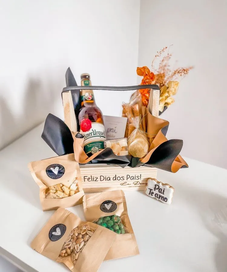 A imagem mostra uma cesta de Dia dos Pais com uma garrafa de bebida, pacotes de petiscos e um cartão. As cores predominantes são marrom e dourado, transmitindo um ar sofisticado. Flores secas adicionam um toque decorativo. O cartão diz “Feliz Dia dos Pais - Pai Te Amo”.