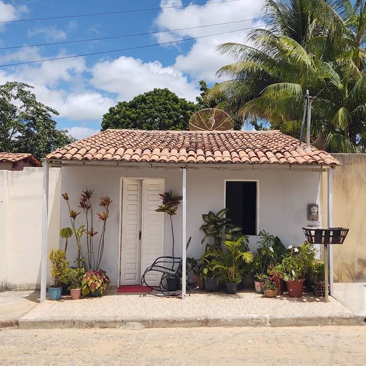 Fachada de casa simples e humilde. Casinha pequena com plantas diversas em vasos e cadeira de balançar