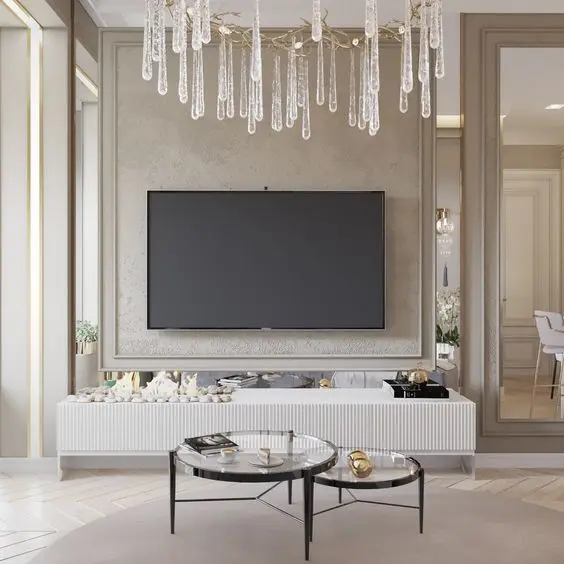 Sala de TV com parede bege com boiserie, lustre, aparador branco com itens decorativos e mesa de centro de vidro 