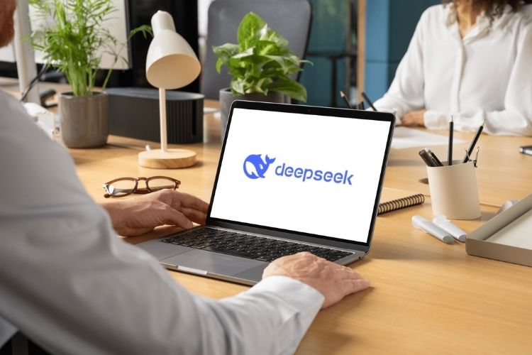 Um homem em um ambiente de escritório observa o logotipo "DeepSeek" exibido na tela de um laptop. A mesa contém itens como óculos, plantas, luminária e cadernos, enquanto outra pessoa ao fundo trabalha em um ambiente organizado.