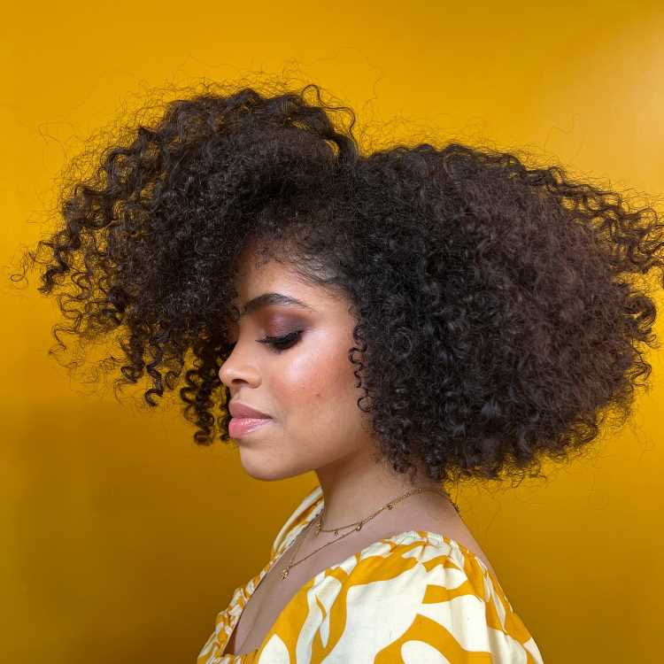 A imagem mostra uma pessoa com cabelo cacheado e volumoso, cortado em camadas repicadas. O corte ajuda a definir os cachos e distribuir o volume de forma equilibrada, criando um visual cheio de movimento. Esse estilo é frequentemente chamado de "corte em camadas repicadas" para cabelos cacheados, ideal para realçar a textura natural dos cachos.


