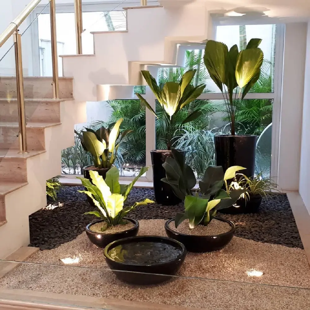 Jardim de inverno embaixo da escada, com pedras claras e escuras, vasos de diferentes tamanhos e muita iluminação