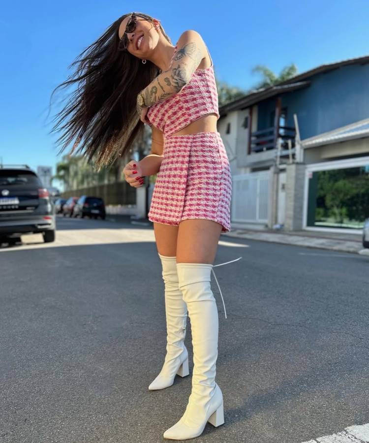 Uma mulher com longos cabelos escuros e tatuagens, sorrindo e com o cabelo ao vento, veste um conjunto rosa e branco de cropped e shorts xadrez, combinado com botas brancas de cano alto, em uma rua ensolarada.