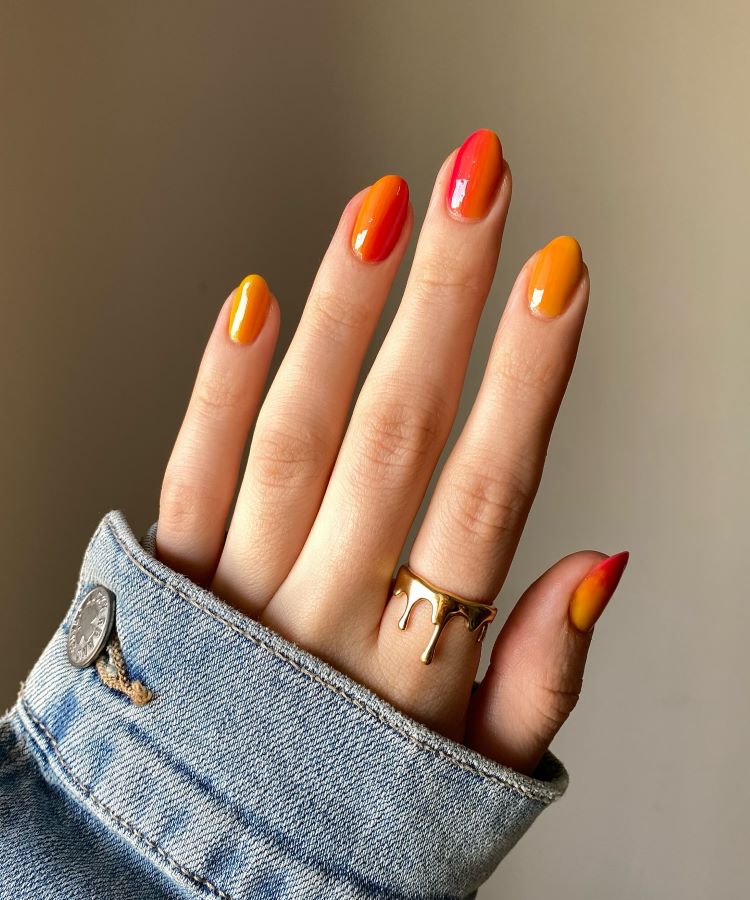 A imagem mostra uma mão com unhas decoradas em um estilo gradiente, variando entre tons de laranja e vermelho. O efeito cria uma transição suave de cores, conferindo um visual vibrante e moderno. A mão está parcialmente coberta por uma jaqueta jeans, e um anel dourado com design fluido adorna um dos dedos. Este estilo é um exemplo de unha decorada simples, que combina criatividade com elegância.
