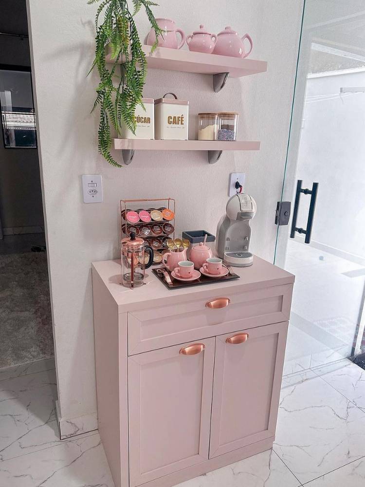 Cantinho do café rosa, com armário, xícaras, prateleiras e decorações em rosa.