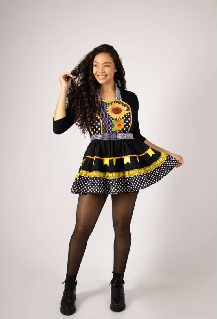 Uma mulher de cabelo escuro cacheado sorri, vestindo um avental junino sobre uma blusa preta. O avental é preto com um grande girassol, babados amarelos, e uma saia de bolinhas brancas. Ela usa meias-calças e botas pretas.


Fontes







Fontes





