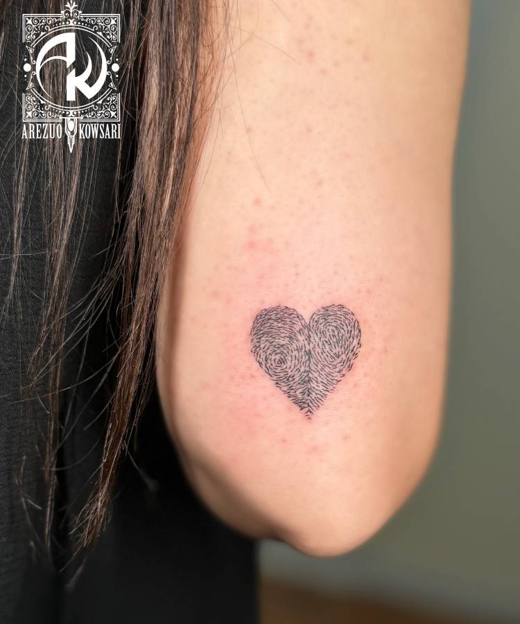 A imagem exibe uma pequena tatuagem de coração no braço de uma pessoa. O coração é preenchido com um padrão que se assemelha a uma impressão digital, em tons de cinza escuro. Parte do cabelo escuro da pessoa é visível. No canto superior esquerdo, há um logotipo e o nome "AREZUO KOWSARI".







