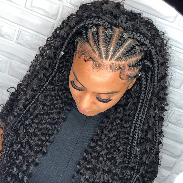 Trança nagô até metade do cabelo em fios cacheados, possivelmente aplique. Mulher está com a cabeça abaixada, usando blusa preta em fundo parede de tijolinhos