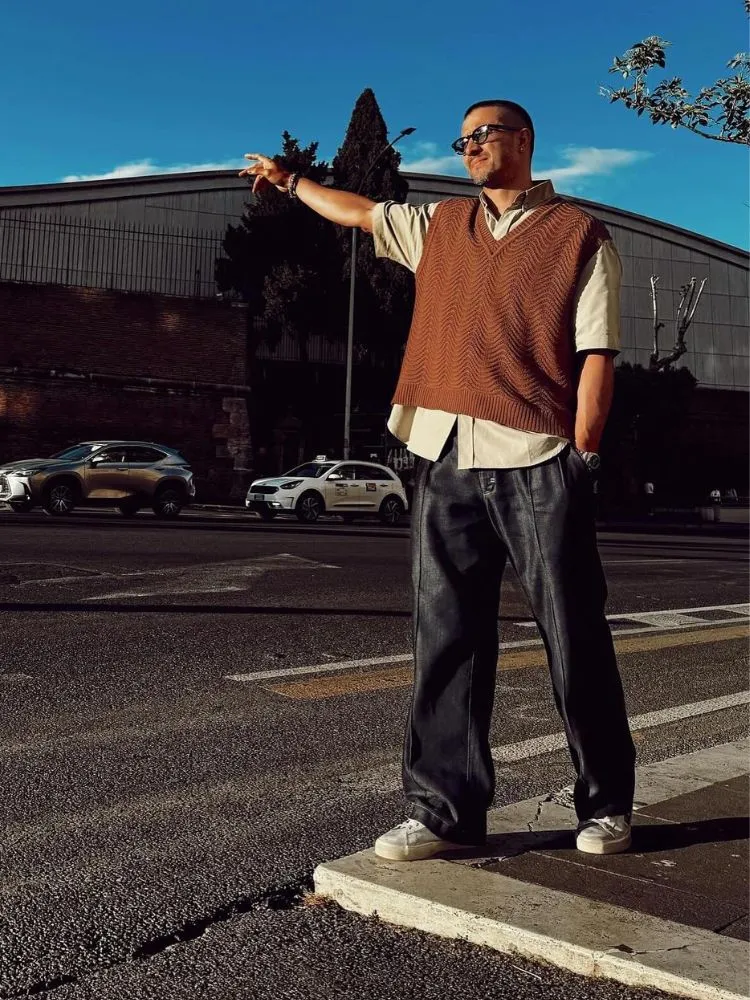 Ator Rômulo estrela, de silhueta masculina trapézio, usando camisa off white, colete marrom e calça jeans escura
