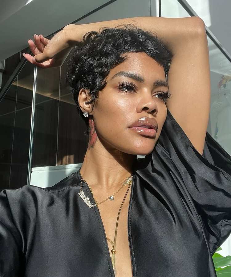 Mulher de cabelo curto cacheado preto, com pele iluminada recebendo luz natural, destacando sobrancelhas delineadas, cílios longos e lábios brilhantes. Usa brinco pequeno brilhante e exibe uma tatuagem no pescoço, posando com expressão confiante.