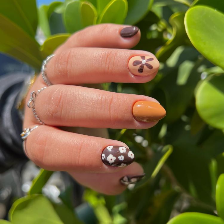 Mão com estilos de unhas curtas, decoradas com esmalte marrom, laranja e nude. Alguns dedos possuem detalhes florais brancos e uma flor estilizada. Fundo com folhas verdes em um ambiente iluminado, destacando o contraste entre as cores vibrantes e naturais.
