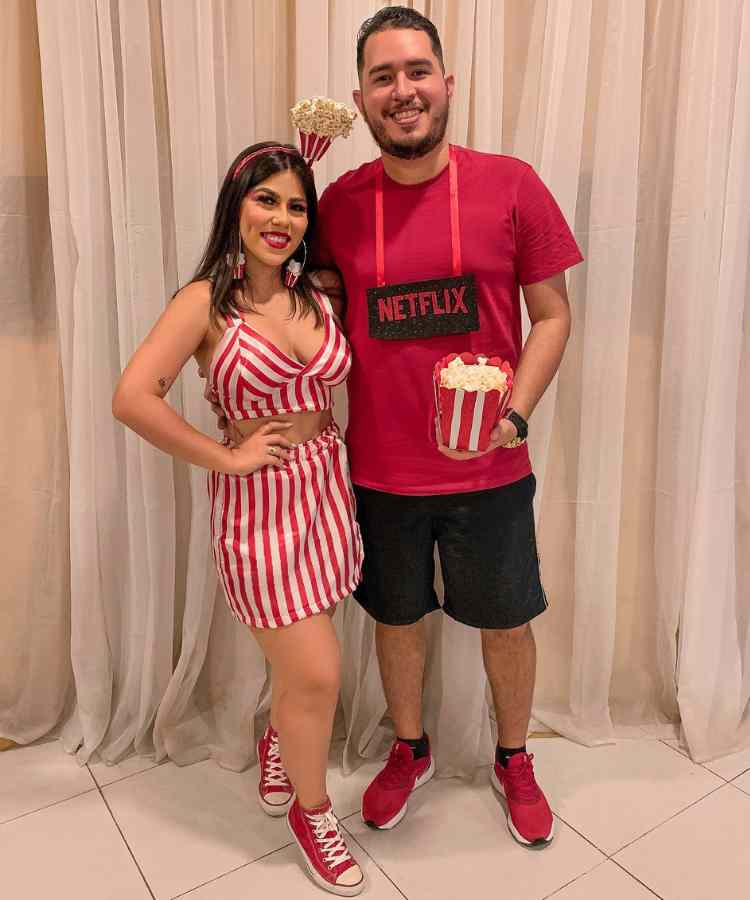 Traje de Dia das Bruxas para namorados: Netflix e Pipoca. Ela usa tiara de pipoca, cropped e saia vermelho e branco. Ele usa plaquinha escrito "netflix", segura pipoca 