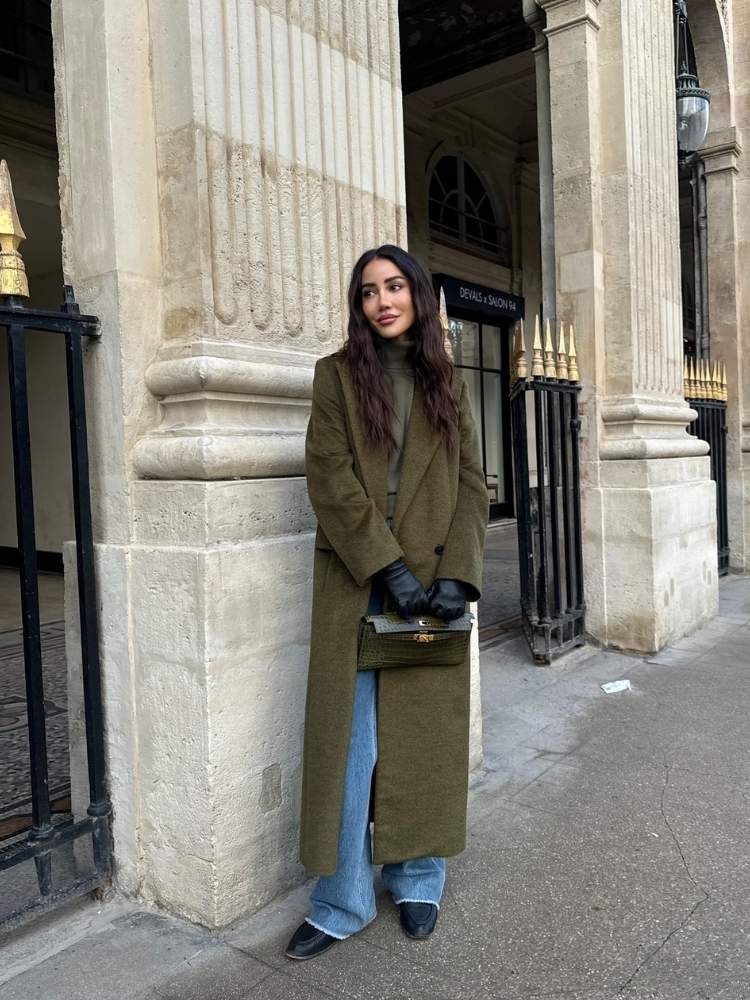 Mulher de pele clara de sobretudo verde-musgo e calça jeans com desgaste na barra