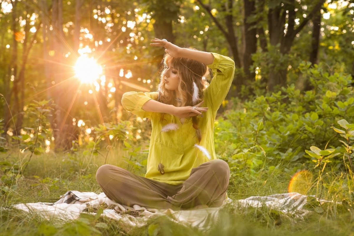 Mulher sentada na natureza ao entardecer, usando roupas em tons de verde, com penas decorativas no cabelo. Ela faz gestos elegantes com as mãos enquanto o sol brilha intensamente entre as árvores ao fundo, criando atmosfera serena.