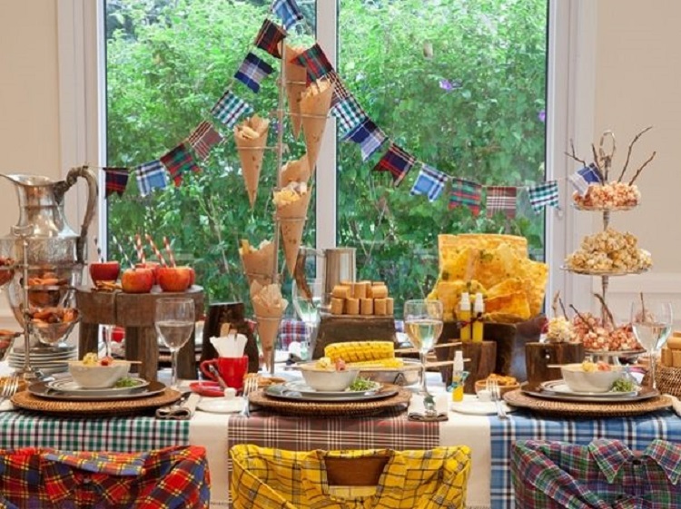 A imagem representa uma decoração de festa junina colorida. Contém mesa com comidas como pipoca, paçoca, pasteis, doces, além de enfeites como bandeirinha, toalha xadrez e camisa xadrez