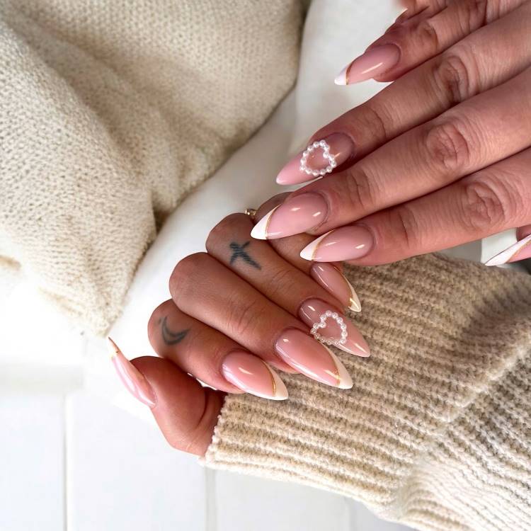 Mãos femininas com unhas longas em formato amendoado, decoradas com esmalte rosa nude, pontas francesas brancas e detalhes em pérolas formando corações. Tatuagens sutis adornam os dedos. Uma malha bege ao fundo cria um cenário aconchegante e elegante.





