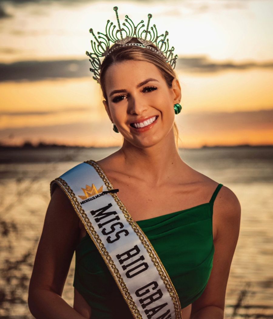 A candidata do Rio Grande do Sul posa com um vestido verde e a faixa de miss, com uma coroa em frente ao mar