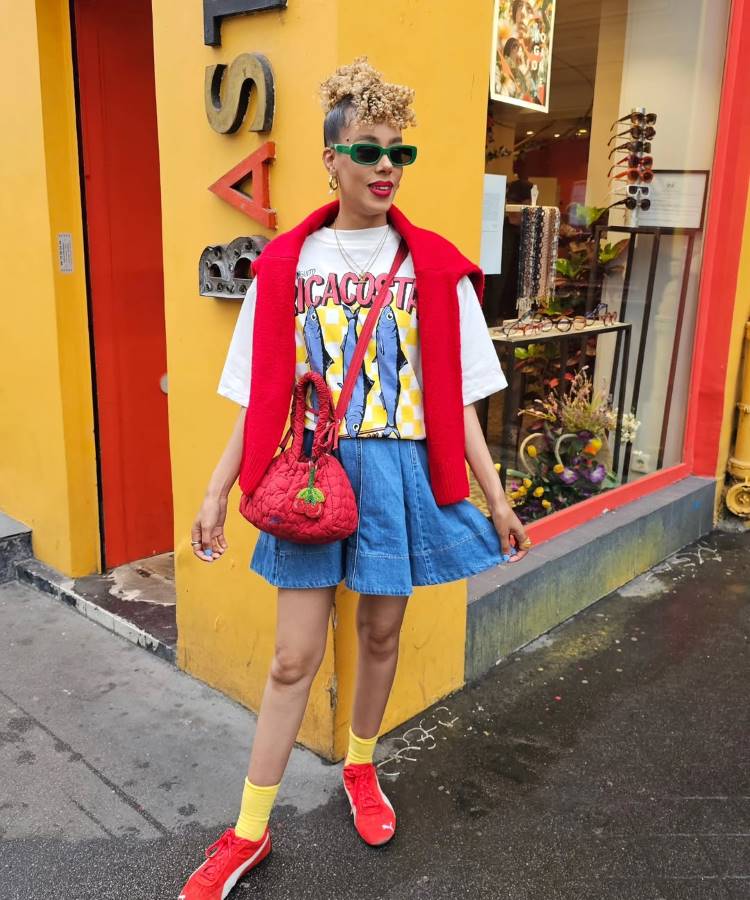 Uma mulher com um coque e óculos de sol verde, usando uma camiseta gráfica, saia jeans e tênis vermelhos. Com um suéter vermelho nos ombros e uma bolsa vermelha, ela exibe as tendências do verão europeu em frente a um prédio amarelo.