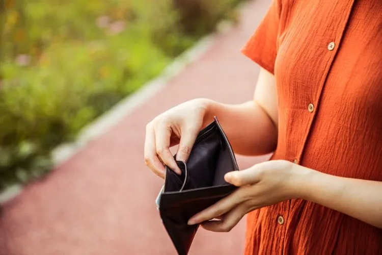 Uma pessoa, vestindo uma camisa laranja, está segurando uma carteira preta aberta e aparentemente vazia. A cena, com um caminho ao fundo, pode ilustrar o desafio de sair do vermelho e a busca por soluções financeiras.

